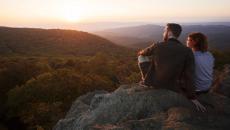 Two serene-looking people sit side-by-side on large rocks overlooking the hills and forests of Shenandoah National Park in Virginia, USA at sunset. The glow of the sun reflects on their faces.