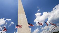 A picture of the Washington Monument from the ground looking up towards the blue, cloud-dotted sky. There are American flags flying around the base of the monument