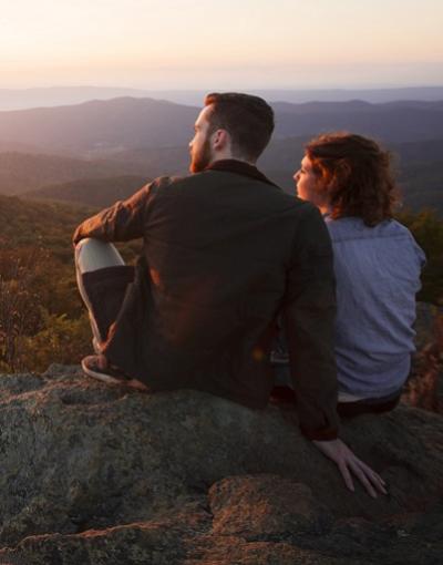 Two serene-looking people sit side-by-side on large rocks overlooking the hills and forests of Shenandoah National Park in Virginia, USA at sunset. The glow of the sun reflects on their faces.